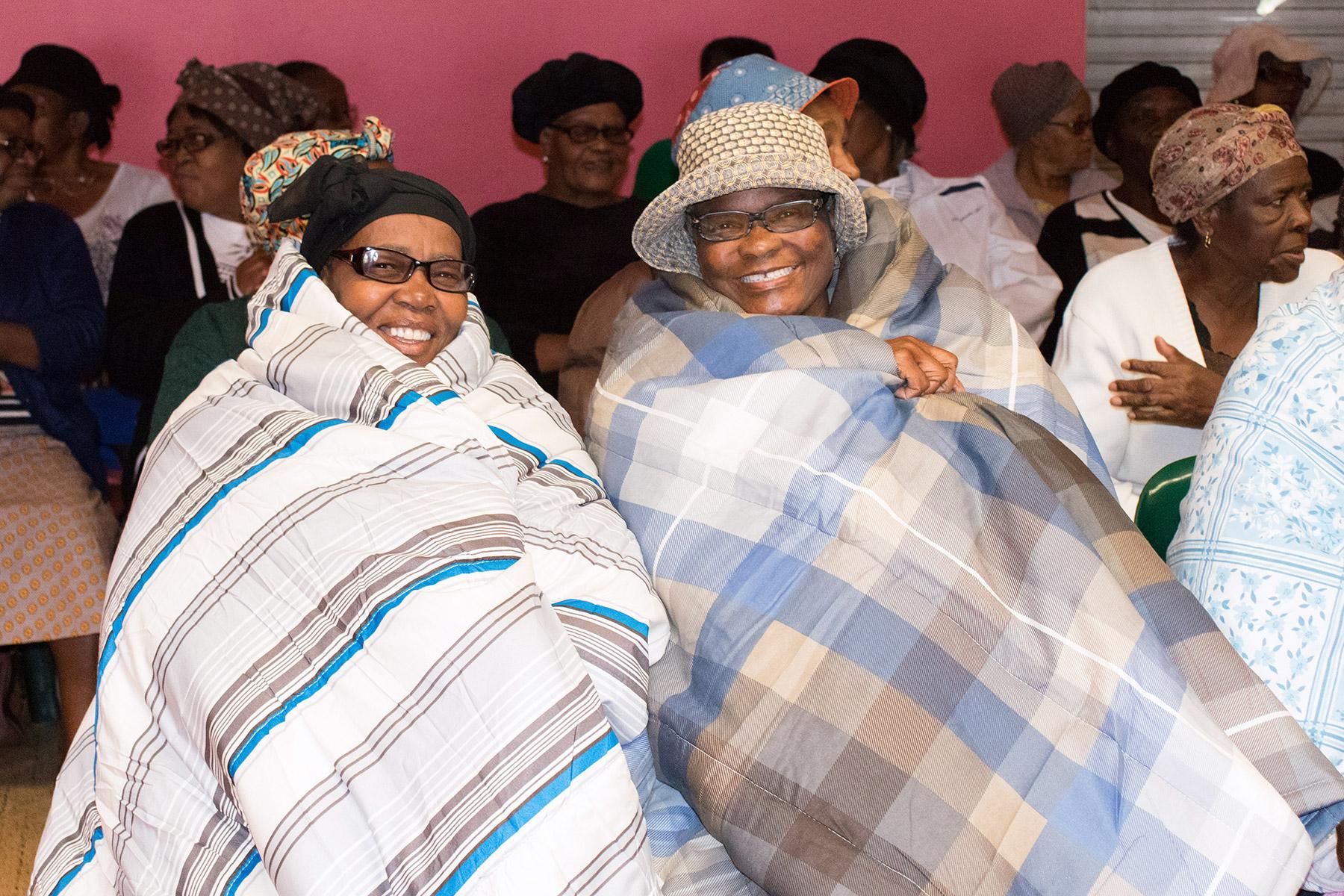 Die älteren Frauen, die sich im Busiswe Luncheon Club, Südafrika, treffen, freuen sich über die Quilts, die sie aus Kanada erhalten haben. Der Treffpunkt bietet ihnen die Gelegenheit, sich täglich zu treffen, um zu singen, gemeinsam zu essen und sich gegenseitig zu unterstützen, während ihre Familien arbeiten und in der Schule sind. Foto: CLWR