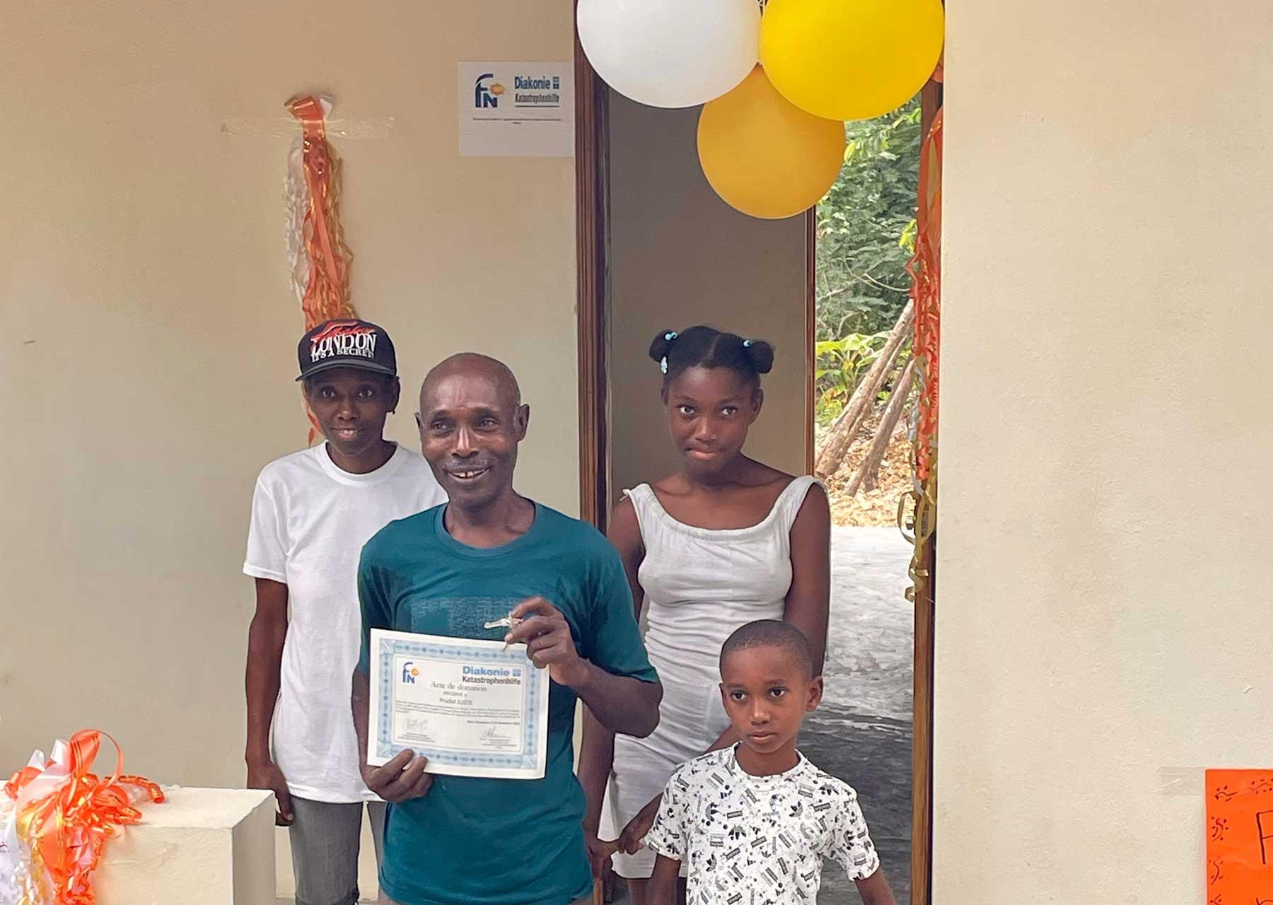The Family Pradel, community of Fon Kochin, Grand Anse, received the key to their new house, built by the Joint Office as part of the response to the August 2021 earthquake. Also pictured is the shelter they have been living in for the past two years. Photo: LWF/ M. French