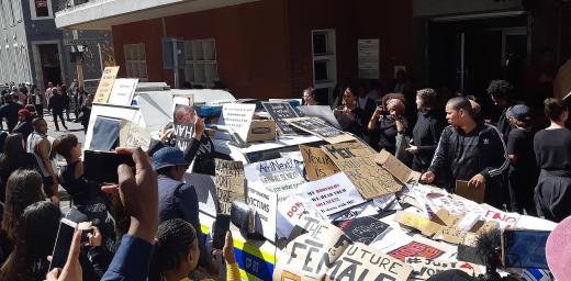 The anti-femicide demonstration in Cape Town, South Africa following the death of Uyinene Mrwetyana. Protestors cover a police vehicle with placards. Photo: Discott, via Wikimedia Commons (CC-BY-SA)